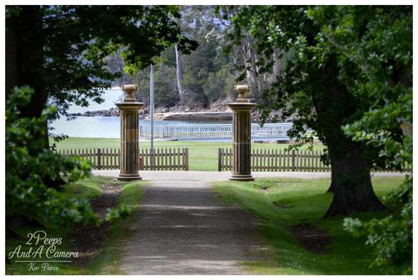 A slightly shaded, gravel path framed by large trees leads through an open gate with two classical stone columns toward a grassy lawn, a picket fence, and the calm blue water of the bay at Port Arthur.

Photo by Kev Peirce.