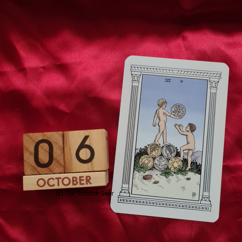 A photo of a wooden block calendar showing the date 06 October next to a tarot card - the 6 of pentacles. The card shows a child standing on top of 5 coins and holding one coin in their hand. Another child has their arms outstretched. It's not clear who is giving and who is receiving.