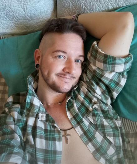 Trans man with brown short hair and facial hair. He has piercing blue eyes, and reclines against pillows. His green, grey and white plaid shirt is open, and he wears a cross around his neck. Be is looking suggestively into the camera, with a slight smile.