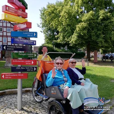 Auf dem Bild sind zwei ältere Damen in einer Fahrradrikscha zu sehen, die von einer jüngeren Frau gefahren wird. Das Gefährt steht neben einem bunten Wegweiser, der verschiedene Ziele anzeigt. Im Hintergrund ist ein Fachwerkhaus zu sehen und Bäume zu sehen.