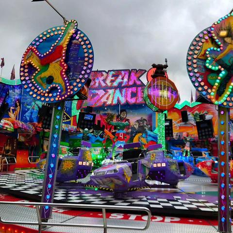 Bunte Jahrmarktsattraktion „Break Dance“ mit grellen Lichtern, bemalten Rückwänden und rotierenden Fahrkabinen vor grauem Himmel.


