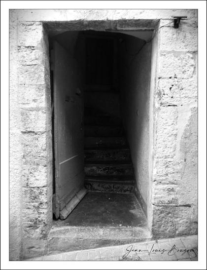 This black-and-white photograph captures a quiet, somewhat mysterious scene: an old stone doorway leading into a narrow, dimly lit stairwell. The texture of the stone surrounding the doorway is rough and weathered, telling a story of age and exposure to the elements. The open door, slightly ajar, invites a glimpse inside but also hints at uncertainty, as the darkness beyond obscures what lies further in.
The stair treads inside the entryway appear worn and uneven, suggesting repeated use over many years. Shadows fall in deep contrast, emphasizing the narrow, confining space and creating a mood that borders on eerie or contemplative. The monochrome palette enhances the sense of timelessness, abstraction, and focus on form and texture rather than color.
Symbolically, doorways often represent transitions or passages between different states or realms—physical or metaphorical—such as moving from the known to the unknown, safety to risk, or one phase of life to another. The partially open door could symbolize invitation, secrecy, or hesitation.
Artistically, the image plays with light and shadow to highlight the architectural detail and the emotional resonance that an ordinary threshold can carry, especially when stripped of color and reduced to light, dark, and texture. The photo connects deeply with themes of mystery, memory, and the stories old structures silently hold.