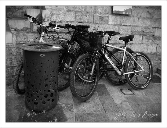 This black-and-white photograph captures an urban scene centered around a small cluster of bicycles parked against a textured stone wall, which adds a rustic and somewhat historical ambiance to the setting. The stone blocks are uneven, giving the backdrop an aged and weathered quality, suggestive of an old town or a historically rich area.
In the foreground, there's a distinctive trash bin with a perforated design. The holes are irregular in size and shape, creating an interesting contrast between the rigid lines of the bicycles and the organic pattern of the bin. The bin’s metal surface seems worn, hinting at frequent use and contributing to the overall textured feel of the scene.
The bicycles themselves appear well-used, equipped with various practical accessories such as baskets and racks, suggesting they are primarily for daily commuting rather than recreational use. The framing of the image focuses closely on the bicycles, allowing for intimate appreciation of their mechanical details—the spokes of the wheels, the chain, and the handlebars all stand out crisply in the monochrome palette.
Artistically, the choice of black and white invites viewers to concentrate on form, contrast, and texture rather than color, evoking a timeless quality. This can symbolize the enduring nature of urban life, where old and new coexist. The bicycles symbolize mobility and freedom, set against the unchanging stone wall, symbolizing stability and history.
