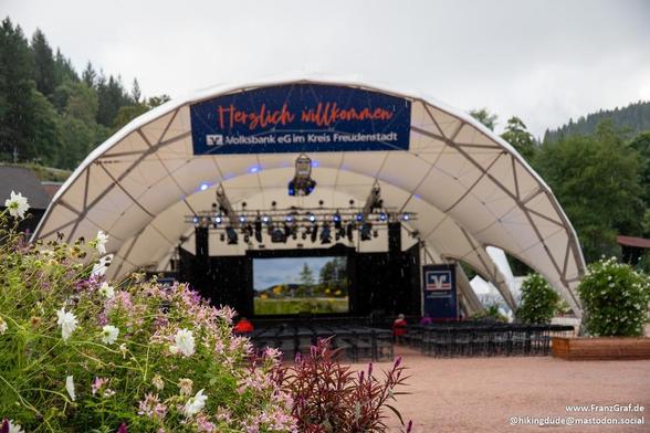 A stage is set up outdoors with rows of white chairs arranged neatly. The stage is illuminated by bright lights, creating a warm and inviting atmosphere. In the background, there is a clear blue sky and tall trees, adding to the natural outdoor setting. A sign is displayed prominently on the stage, catching the eye with its orange writing against a blue background. The overall scene exudes a sense of preparation for a performance or event, with the stage ready for speakers or performers to take…