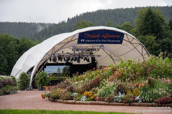 A serene outdoor scene featuring a white tent with a stage set up in a garden filled with colorful flowers. The tent is illuminated with lights, creating a magical ambiance. Surrounding the tent are various types of plants and trees, including one with white flowers. The sky above is clear with a few fluffy clouds, adding to the picturesque setting. The overall color scheme is dominated by white and grey tones, with accents of a deep green color. The atmosphere suggests a festive event or perfo…