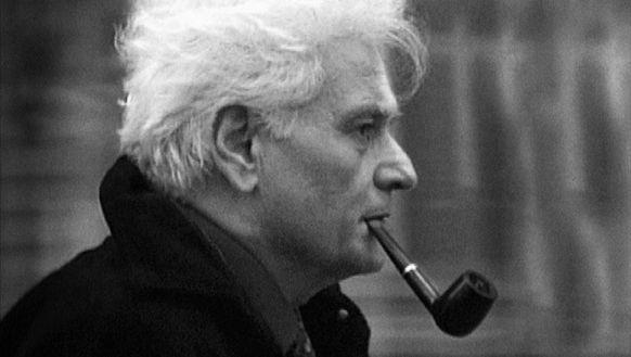 Jacques Derrida, philosophe français connu pour sa théorie de la déconstruction. Il est vu de profil, avec ses cheveux blancs et il fume une pipe. Il porte un manteau sombre.