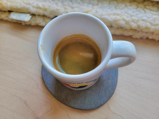 A white, 3 ounce, ceramic espresso (demitasse) cup with a shot of espresso, on a gray diatomaceous-earth coaster on a maple desktop. The blue text on the cup reads "Cyber-Physical Systems Research Center" and wraps in a circle around the logo, which looks like a loosely interlaced, rounded triquetra with thick knots in solid red, yellow, and blue (each its own color). Next to the cup is the edge of a matted sherpa-fleece blanket.