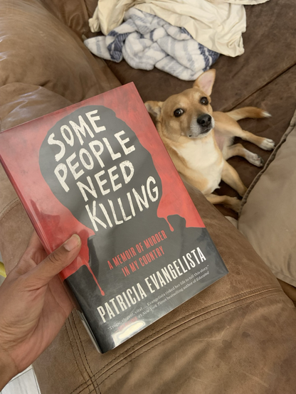 The cover of the book Some People Need Killing displays a blacked out silhouette of Duterte against a red background. The title is in all caps seemingly hand-written. "A memoir of murder in my country is in capitalized red subtext above the name of the author also in an all caps white text.    The book is being held above a tan couch on which lies a small light brown dog that is looking up at the camera. 