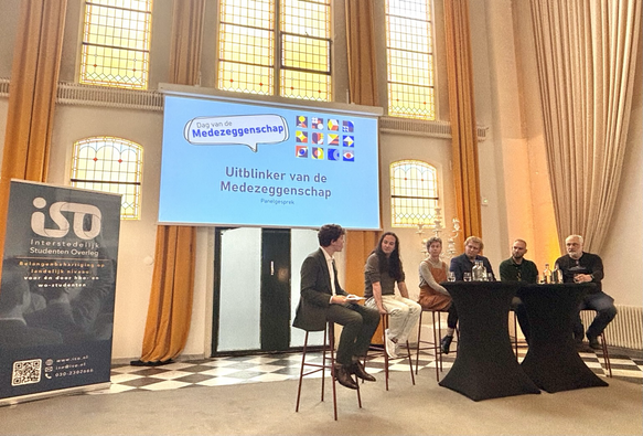 Foto van een grote zaal met hoge oranjeachtige gordijnen en geel licht door de ramen. Links een blauwe banner van het ISO (interstedelijk studenten overleg). In het midden een groot scherm met blauwe achtergrond en tekst “uitblinker van de medezeggenschap” en rechts daaronder 6 mensen op barkrukken aan twee sta-tafels die in gesprek zijn. 