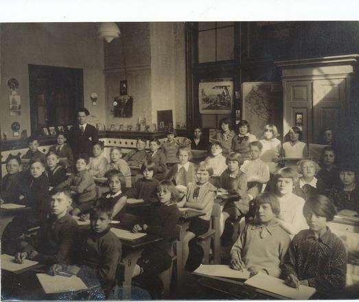 Do you treat conference attendees as adults? Black and white photograph of a 1940s classroom full of children sitting at desks and looking forward. A teacher, wearing a suit, stands at the back of the room. Photo by Flickr user yvonnert