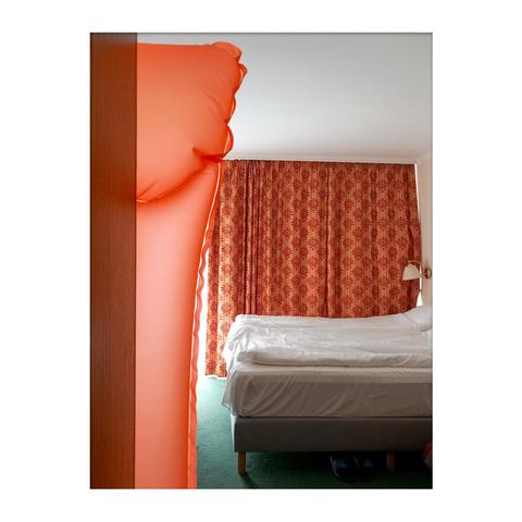 The picture shows a hotel room. In the foreground of the picture, there is an orange rubber mattress propped against the wall. In the middle of the room is a double bed, a reading lamp on the wall, and in the background, an orange blackout curtain is drawn in front of the window, which rhymes nicely with the rubber mattress in the foreground in both form and color.