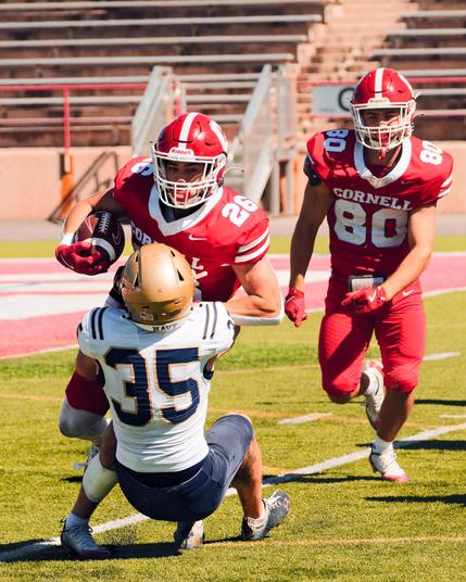 On the the left side number 35 in white jersey and black pants is hanging on to the front of 26 in red as 26 holds the football,  to the right red 80 is running foreward,  behind that action the end zone and a set of empty bleachers