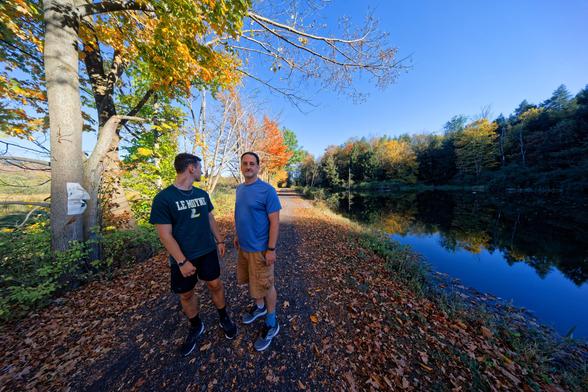 One side of a trail runs right down the center of the frame,  to the left there are two men,  leftmost wearing a green shirt that say LE MOYNE with an L logo is looking to the back while the rightmost is wearing a blue shirt and lookig forward,  the left is a narrow hedgerow and then the open area of Dryden Lake,  to the right a pond lines the trail and in the far distance can be seen orange and green trees.