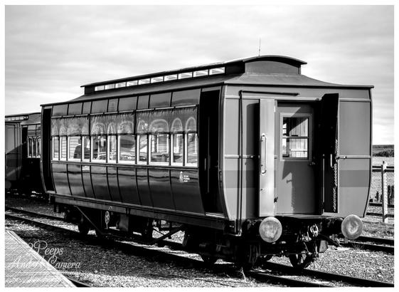 A striking black and white photograph of an old, preserved passenger train carriage, possibly from the Abt Railway, sitting on the tracks at Strahan, Tasmania.

The carriage features large windows along its side and open doors at the end. The image, signed Kev Peirce Photography, captures the vintage rail heritage under an overcast sky.