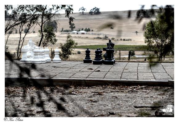 Giant white and black chess pieces (Kings, Queens, Bishops, Knights, Rooks, and Pawns) are set up on an outdoor, oversized wooden chessboard.

The black pieces are on the right, and the white on the left. The board sits on a gravel and dirt ground.

In the blurry background, a dry, rolling hill and a distant farmhouse are visible under a bright sky.

The scene is slightly framed by dark, out-of-focus foliage in the foreground. Photo by Kev Peirce.