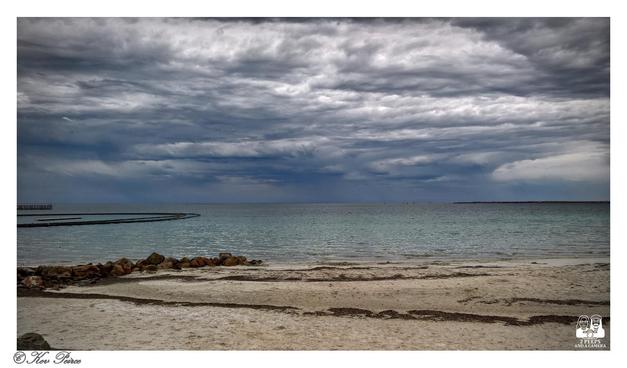 A wide angle landscape photograph of Wallaroo Beach under a heavy, dramatic storm sky.

Dark, textured grey clouds fill the top two thirds of the frame, with a lighter, ominous break of rain visible on the right horizon.

The middle ground is the calm, turquoise-grey ocean. In the foreground, there is a stretch of light tan sand with patches of dark seaweed, and a small pile of dark brown rocks on the left.

A curved, dark structure, possibly a breakwater or netting, extends into the water from the left.