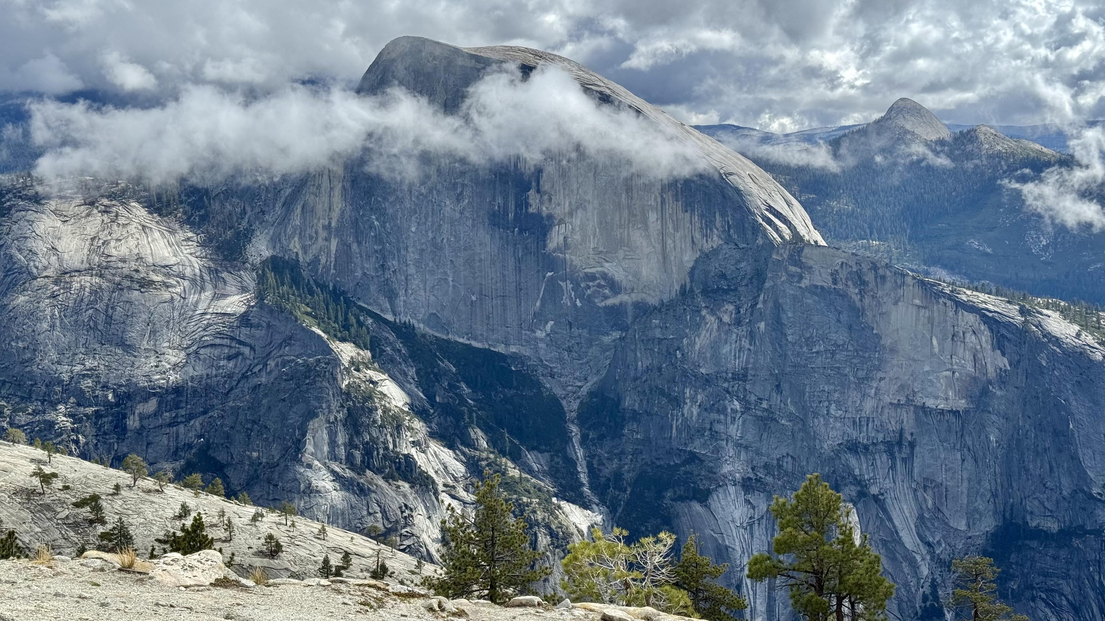 Half Dome from the North Dome trail, clouds lingering. Some scattered trees on the nearer granite.