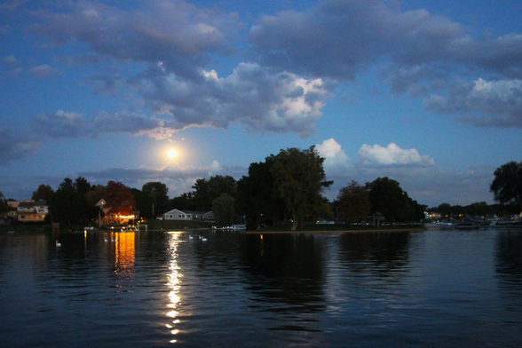 Photograph of a full moon in a cloudy blue sky with a lake in the foreground.
