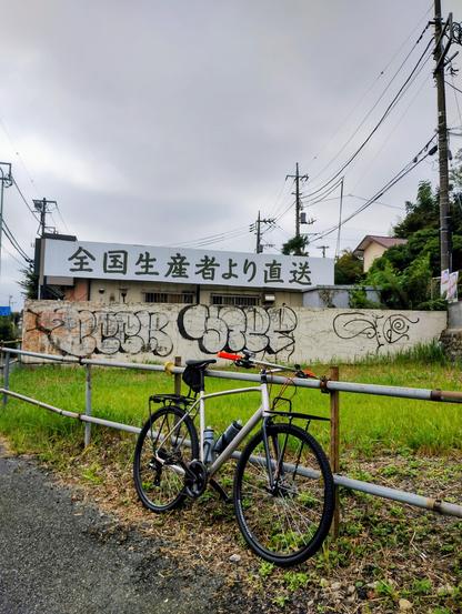 A silver flat bar bike in front of a scaffold fence, graffiti wall & a Japanese sign advertising direct from the farm produce.