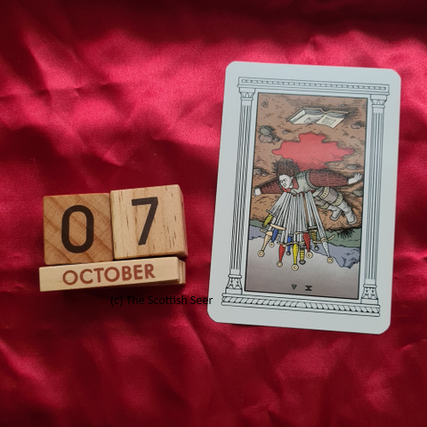 A photo of a wooden block calendar showing the date 07 October next to a tarot card - the 10 of swords reversed. The card shows an adult lying on the ground, on their back, in a pool of blood. They have 10 swords impaled through their chest.
