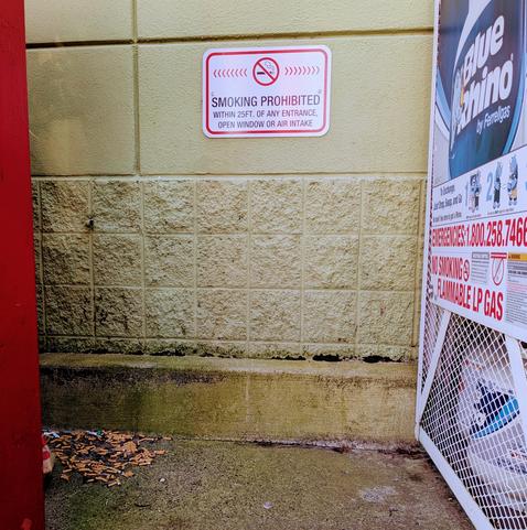A concrete nook with a horde of cigarettes butts in a corner, near a sign that says "SMOKING PROHIBITED within 25 ft of an entrance, open window, or air intake". On the right there are stacks of propane canisters in a metal cage that also has "NO SMOKING" in prominent red letters. The propane brand is Blue Rhino. The propane founder Billy Rhim imagined the Blue Rhino to be the Pink Panther's roommate in an odd couple situation, but less Neil Simon and more Clive Barker. Billy himself wrote thou…