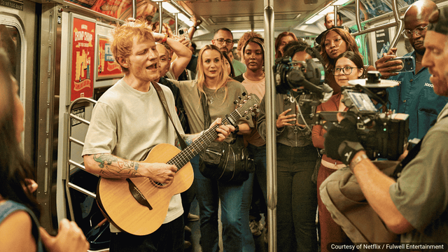 Ed Sheeran stands inside a New York City subway car holding a guitar, singing to a small crowd of passengers who listen and smile as the train moves. Scene from One Shot with Ed Sheeran, Netflix’s one-take music special premiering Nov. 21, 2025. | Image: Courtesy of Netflix / Fulwell Entertainment