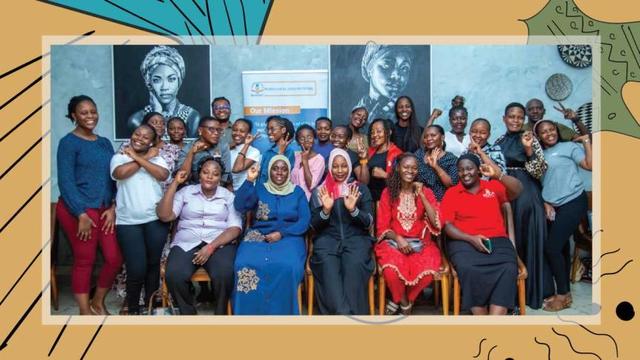 Group photo features approximately 30 women of African descent, gathered in a workshop organised by WOUGNET. Most of the women are standing, with a few seated in the front row, all smiling and engaging with the camera; some are raising their fists or hands in gestures of solidarity or enthusiasm. They are dressed in a variety of colorful clothing. In the background, there's a wall adorned with two large black and white portraits of women, and a banner or sign with text that includes "Our Mission." The overall image is framed with a decorative, abstract border in shades of beige, teal, and black, adding a stylistic touch.