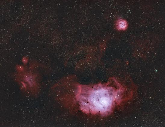 This image is a photograph of the Lagoon and Trifid Nebulae, located in the dense core of the Milky Way. Both celestial bodies appear as bright, gaseous shapes, rendered in shades of pink, red, and blue. Surrounding the nebulae are bright pinpricks of light, which represent stars. 