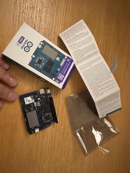 An Arduino board is placed on a wooden surface, next to its packaging, an instruction manual, and a small plastic bag. And Lucy's fingers. The fingers are still attached to Lucy, but she's not in shot. 