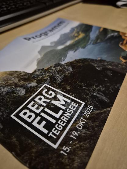 A close-up of an eye-catching program brochure for the **Bergfilm Tegernsee** event, held from October 15 to 19, 2025. The brochure features a striking cover with a rugged mountain landscape and a serene alpine lake, evoking a sense of adventure and natural beauty. The bold, modern typography of the event name stands out, inviting outdoor and film enthusiasts to explore inspiring stories of mountain adventures and landscapes.