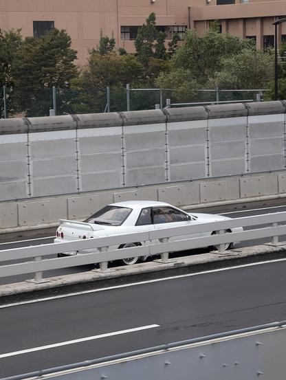 A white sports car with its lower half hidden by a guard rail drives along a tokyo expressway. 
