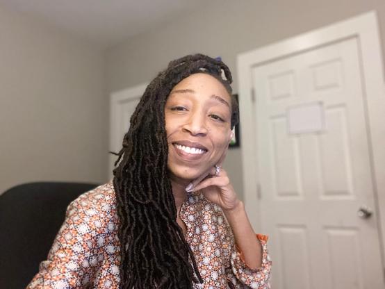 Sporting long faux locs, The Ratchet Sage™ sits in a softly lit room, resting her chin on her hand. She wears a patterned blouse in warm tones and looks calm, confident, and grounded. The gentle light and relaxed pose capture the theme of strength through stillness — the quiet resilience of recovery and sovereignty.