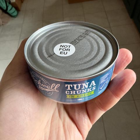 A person holds a can of tuna chunks in brine. The can has a label indicating it's not for the European Union. The background shows tiled flooring.