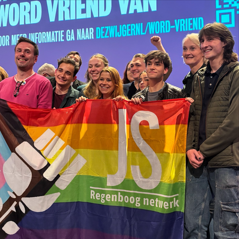 Groepsfoto op het podium, met een grote Pride-vlag (dus de regenboogkleuren + de kleuren voor trans en a-seksualiteit) van de Jonge Socialisten met witte tekst JS Regenboog Netwerk.