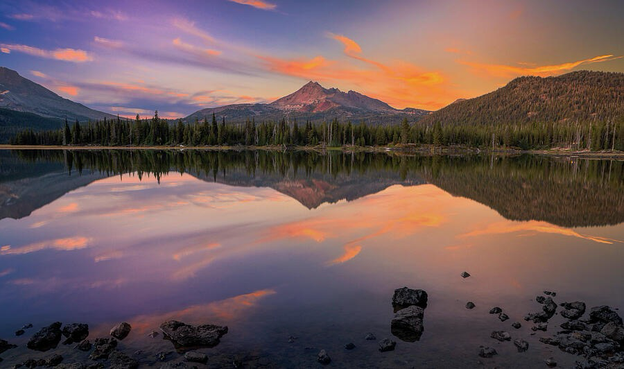 A tranquil view of Sparks Lake in Oregon at sunset. The still water reflects the vivid orange and purple sky as the distant peaks of the Three Sisters rise on the horizon, surrounded by evergreen trees and fading evening light.
