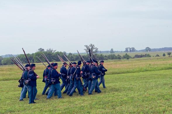 Union Soldiers walking away with rifles on the ancient battlefield on a patch of short-cropped grass with moderate cropped grass and hedgerows in the distance.