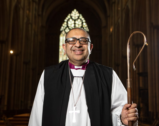 Angliciab Bishop has a purple collar and a silver cross and is wearing glasses and holds a staff like the hook you'd use on a sheep and stands in the middle of a cathedral with stained glass behind
