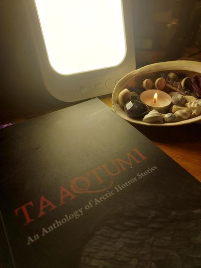 In the foreground is a copy of Taaqtumi: An Anthology of Arctic Horror Stories (ed. Neil Christopher), which features a raven covering the whole of the front and the title in red. Next to the boom is a large sea shell filled with bits of natural materials and a lit tea light. Behind the book and shell is a small, rectangular SAD lamp lit up.