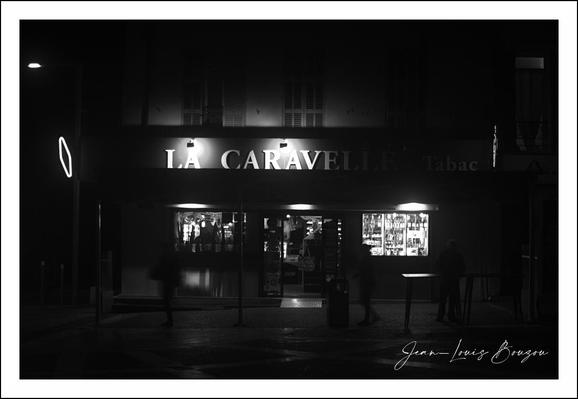 The image is a black-and-white photograph capturing a nighttime street scene with a prominent focus on a storefront illuminated from within. The store's sign reads "LA CARAVELLE," suggesting a likely French establishment, possibly a café or tabac, as hinted by the faint word "Tabac" on the right side. The glow from inside creates a contrast against the dark exterior and the surrounding night, providing a warm, inviting focal point.

Two figures appear outside the shop, one slightly blurred, likely from motion, walking towards or past the entrance, while the other stands more still, shadowed against the lighted window. This dynamic adds a subtle narrative element to the image—movement versus stillness, presence versus absence.

The overall ambiance evokes a quiet, perhaps contemplative moment in an urban setting, with the monochrome palette enhancing the moodiness and timelessness of the scene. The use of black and white strips away the distraction of color, focusing attention on light, shadow, and form. This can also hint at nostalgia or a classic, cinematic feel, often associated with Parisian street photography.

Artistically, "La Caravelle" as a name could symbolically reflect journeys or exploration tying into the theme of travel or discovery, perhaps metaphorically through the stories and interactions within the café itself. The photo captures not just a place but a fleeting moment in urban life, rich with atmosphere and subtle storytelling.