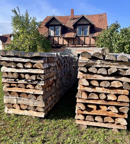 Zwei sehr ordentlich gestapelte Holzpolter aus gespaltenen Meterstücken in einem Garten mit dahinter zwei Obstbäumen und einem Fachwerkhaus.