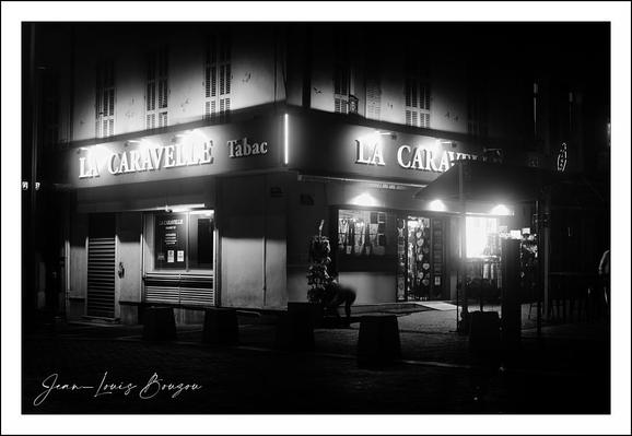 The image presents a nighttime street scene, captured in black and white, imbuing it with a classic, timeless quality. The focal point is a corner building, evidently a café or bar named "La Caravelle," with its illuminated signs starkly contrasting against the dark surroundings. The light from the signs casts a glow that highlights certain architectural details—the windows, doorframes, and parts of the street—and creates deep shadows around the rest, adding a mysterious or even nostalgic atmosphere.
The street outside appears quiet, empty, and still, with an absence of people contributing to a contemplative or solitary mood. The black and white palette emphasizes contrasts and textures—shiny surfaces, stone, or concrete details—while lending a somewhat poetic or cinematic feel reminiscent of film noir. The overall composition is anchored by strong verticals and angles, notably the corner of the building, giving the image a sense of stability amidst the quiet darkness.
This scene could symbolize urban solitude or the beauty found in everyday, overlooked moments. The café itself, often a social hub, stands silent and closed, perhaps suggesting themes of pause or waiting. The aesthetic choices reflect a reverence for light and shadow interplay, evoking feelings of calm introspection or a quiet night’s vigil in a city.
