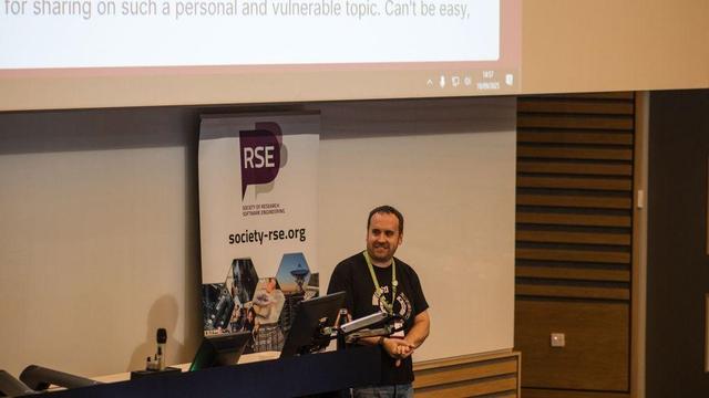 Mike Simpson looking nervous during a talk at the 2025 UK RSE Conference. He's standing at the front of the room next to a lecturn, wearing a black SSI t-shirt and green Mental Health First Aider lanyard.

Photo by Squire & Squire Photography.