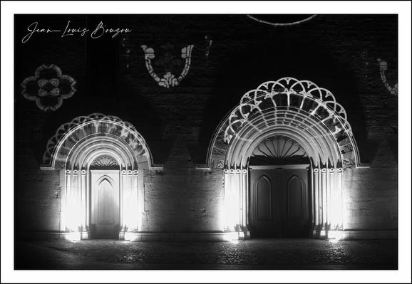 This black and white image captures two intricately illuminated arches set into a stone facade, evoking a sense of historical and architectural grandeur. The lighting accentuates the delicate, lace-like stonework that frames each doorway — the left arch is smaller with concentric, semi-circular layers of intricately carved stone, while the right arch is larger, featuring an elaborate fan-shaped pattern that creates a dramatic play of light and shadow.

Between the two arches, embedded in the wall, there is a small, serpentine ornamental design reminiscent of a horseshoe or stylized "U," which adds an unexpected yet charming accent. Additionally, a floral motif is visible on the left side, enhancing the decorative richness of the scene.

The overall atmosphere is almost theatrical, with the illumination spotlighting the architectural details against the dark backdrop, lending the image a timeless, almost sacred quality. This style of arches and stone detailing might hint at a Romanesque or Gothic architectural influence, often found in old European churches or castles, reflecting cultural heritage and artistic craftsmanship rooted in medieval history.
