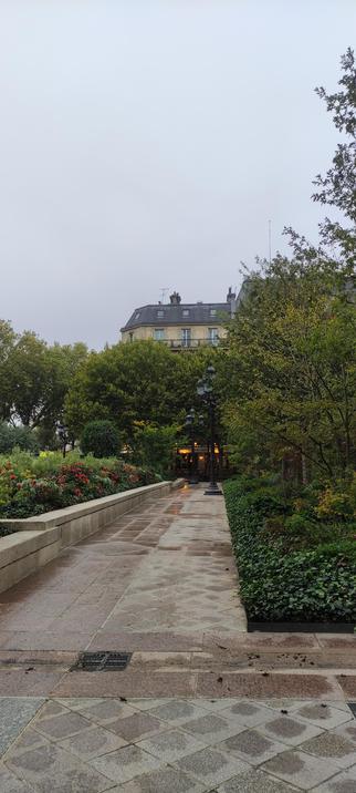 Allée parisienne mouillée par la pluie, bordée de rosiers rouges et de lierre vert sombre. Au bout du chemin, un kiosque éclairé se devine sous les arbres, devant un immeuble haussmannien aux fenêtres encore closes