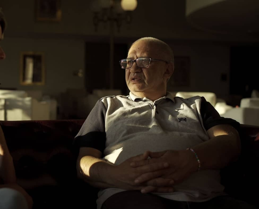 Man with glasses sitting on couch in a white shirt looking to the side as shadows are reflected on his face
