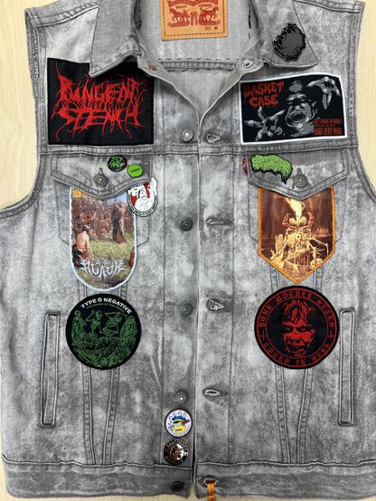 The front of a gray denim battle jacket. 6 metal band and horror patches are visible along with a handful of pins. 