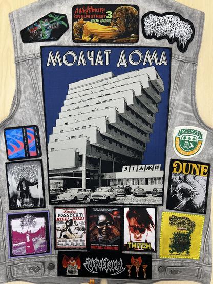 The back of a gray denim battle jacket, mostly covered. Patches include a large Molchat Doma back patch, band patches, and some from horror/exploitation flicks. A Mickey’s fine malt liquor patch is also visible. 