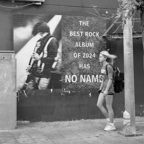A black and white film photo. A woman, with hair flowing in the wind, walks by a poster of a man playing a guitar with hair moving around in Austin, Texas. Tuesday, December 17, 2024.