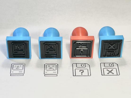 Four plastic stamp handles (three in light blue, one in red) laying on the side showing the bottom of the stamps in black. Below is white paper with four stamps of Classic Mac icons in black ink of the happy Mac, sad Mac, disk with question mark, and disk with an X.
