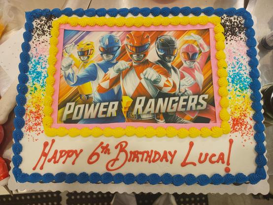 A sheet cake decorated with a Power Rangers edible image and red writing that reads "Happy 6th Birthday Luca!"

The borders reference the other 4 original Ranger colors (black, blue, pink, and yellow), as do glitter flakes on either side of the image. Unfortunately, there is no green decoration due to the lack of the Green Ranger in the image; it's only the initial five characters.